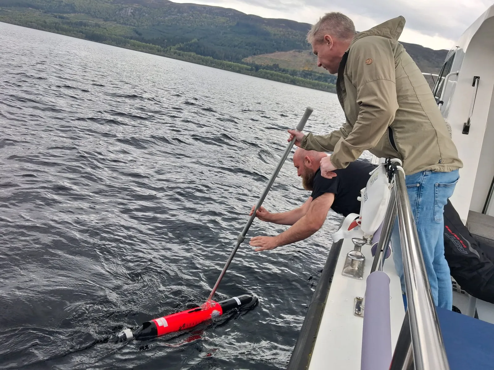 Team members on a boat guiding a red AUV alongside the hull on Loch Ness